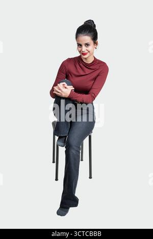 Young woman looking at camera, sitting confidently on a stool in a studio Stock Photo