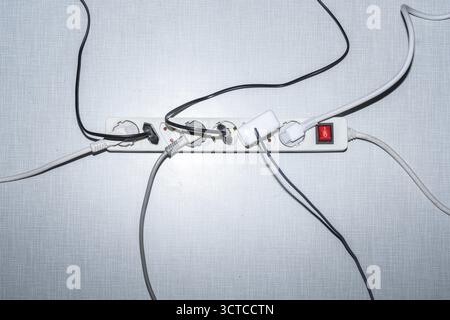 Top view of a power strip with on/off switch and various plugged-in plugs and cables on a white Resopal panel Stock Photo
