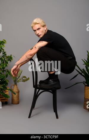 Young man showcases unique fashion while sitting on a chair amidst lush plants. Stock Photo