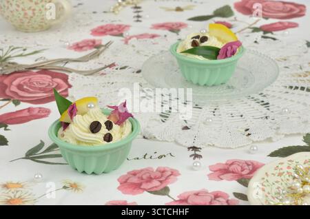 Two decorated, cream cupcakes on a retro tablecloth with flowers, roses and white, crocheted doilies. Painted ceramic and pearl decorations. Stock Photo