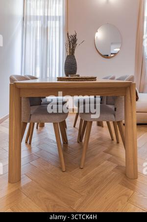 Еру wooden dining table and fabric chairs in a small apartment interior. Features herringbone parquet floor and lavender decor in neutral tones Stock Photo