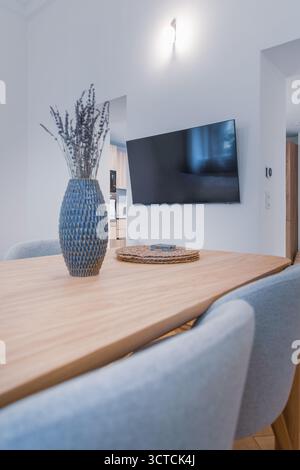 Close-up of a wooden dining table and fabric chairs in a small apartment interior. Features lavender decor and a large wall-mounted TV Stock Photo