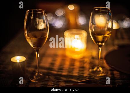 Soft bokeh surrounds twin glasses of white wine on a table lit by flickering candles. The golden light enhances the intimate setting for a special eve Stock Photo
