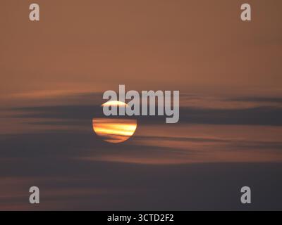 Close-Up Sunset Through Dramatic Cloud Layers - Telephoto close-up of the vibrant setting sun partially obscured by horizontal, smoky clouds, creating Stock Photo