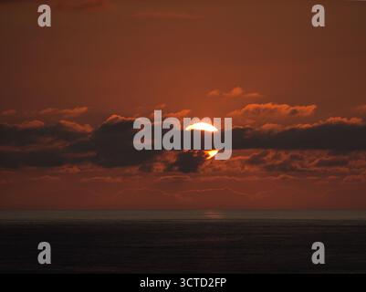Burning Orange Sunset Over Ocean Horizon - Intense telephoto close-up of the setting sun partially hidden by dark horizontal clouds, bathing the ocean Stock Photo