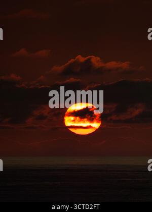 Burning Orange Sunset Over Ocean Horizon - Intense telephoto close-up of the setting sun partially hidden by dark horizontal clouds, bathing the ocean Stock Photo