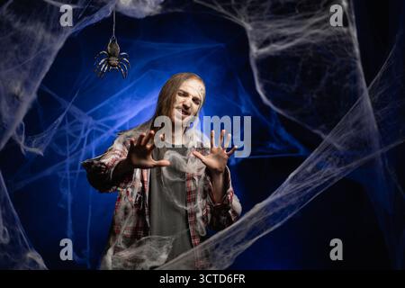 Creepy scary zombie man with ugly makeup holding palms in stop no reject gesture looking at camera standing on spooky spider web background. Halloween Stock Photo