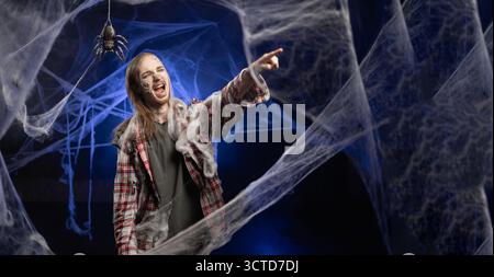 Dead guy with horrible scary Halloween zombie makeup on face pointing fingers, I choose you concept, advertisement while standing covered in spiderweb Stock Photo