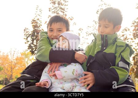 Candid portrait of three siblings, East Asian boys and toddler girl, sitting outdoors during bright fall weather. for family, childhood, and seasonal Stock Photo