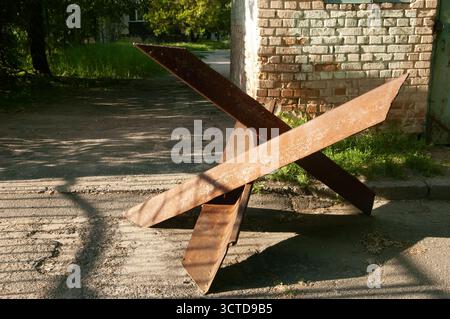 Broken asphalt road with a large rusty tank trap (hedgehog) as the main focus. Behind it, a ruined brick wall, path, grass, and trees in the backgroun Stock Photo