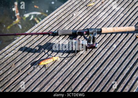 Modern fishing rod with fixed spool reel and lure set up on a wooden jetty by the river, symbolizing outdoor sport and angling lifestyle. Stock Photo