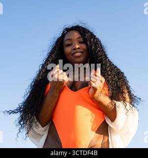 Stunning dark-skinned woman with flowing black curls relaxes at the beach in a vibrant orange swimsuit and white shirt, soaking up the warm weather Stock Photo