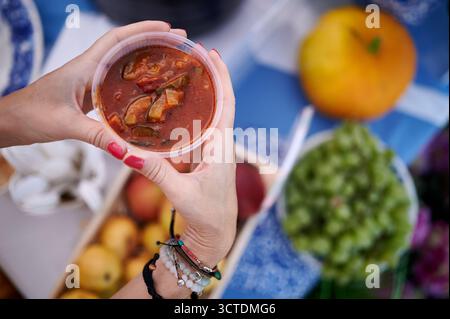 Close-up of tomato soup served in plastic cup above harvest market table with fruits, top-down composition, warm color palette, bright daylight. Stock Photo