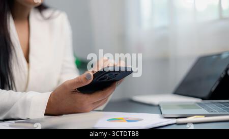 Close-up of businesswoman using smartphone at desk with laptop and financial documents. Concept of digital business, technology communication, remote Stock Photo