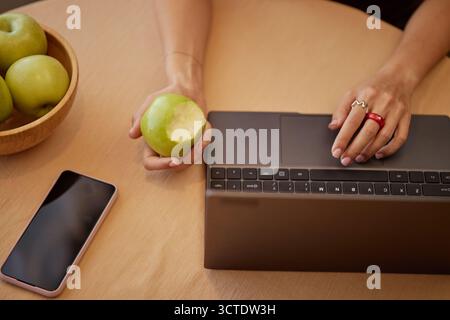 Young Adult Woman Working on Laptop While Eating Green Apple Stock Photo