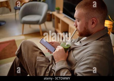 Young Adult Man Sitting Indoors Using Smartphone While Drinking Smoothie Stock Photo