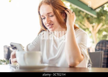A young Caucasian woman with long red hair sits at a cafe table, smiling while using her smartphone. A cup of coffee is placed in front of her. Stock Photo