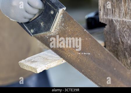 A close up of hand saw cutting through piece of wood, with gloved hand holding saw. image captures texture of wood and rust on saw blade Stock Photo
