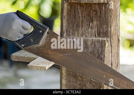 A hand saw is being used to cut wooden plank outdoors, showcasing close up of tool and wood. person is wearing glove for safety, emphasizing focus on Stock Photo