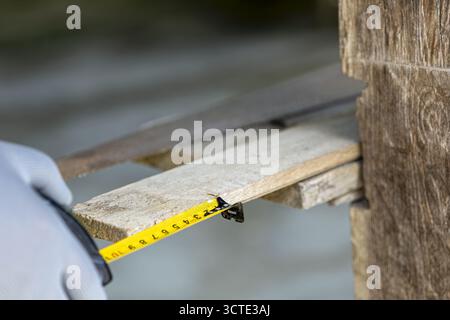 A person wearing gloves measures wooden plank with tape measure, focusing on precision and accuracy in woodworking scene suggests workshop environment Stock Photo