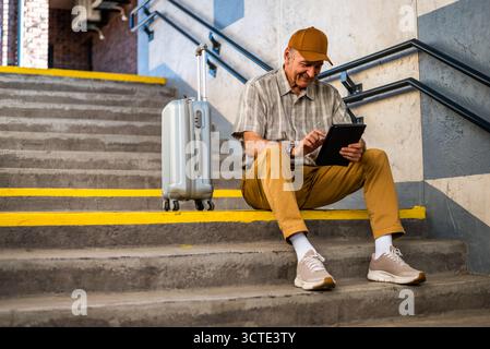 Happy senior man is sitting at railway station and waiting for boarding. He is using digital tablet. Senior people traveling. Stock Photo