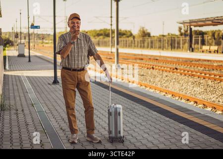 Happy senior man is standing at railway station and talking on smartphone. Senior people traveling. Stock Photo