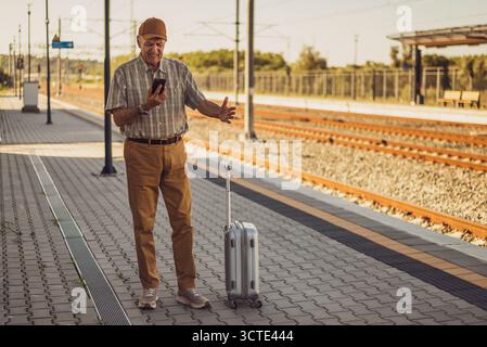 Frustrated senior man is standing at railway station and waiting for arrival of the train. Train departure is delayed. Senior people traveling. Stock Photo
