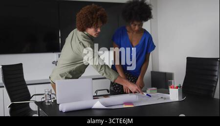 Leaning over rolled-out blueprint two colleagues collaborating at table, with laptop, sticky notes Stock Photo