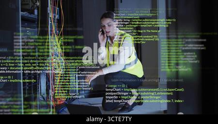 Kneeling IT tech in hi-vis vest configuring network cables in server room with smartphone, laptop Stock Photo