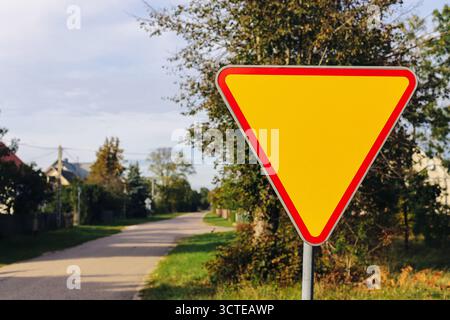 Triangular yield road sign on countryside street with clear sky. Traffic control and road safety symbol in rural area with trees and houses. Stock Photo