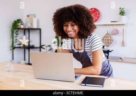 Happy young multiracial woman in apron using laptop while cooking in the kitchen Stock Photo