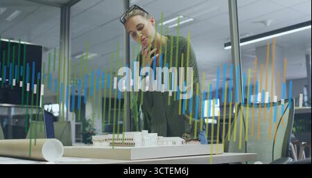 Leaning architect examining scale model at design studio, with blueprint tube laptop and eyeglasses Stock Photo