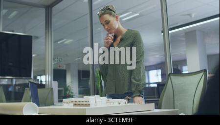Examining woman reviewing white scale model in open-plan office, with laptop and rolled blueprints Stock Photo