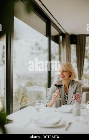 A woman sits at a elegantly set table in a bright restaurant, holding a glass of red wine while looking out at a beautiful outdoor view. Sunlight stre Stock Photo