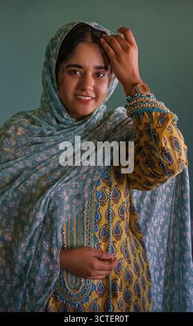 Isfahan, Iran - 01 May 2025: View of a woman in a patterned yellow and blue dress and headscarf, her smile illuminated against the soft turquoise backdrop. Stock Photo