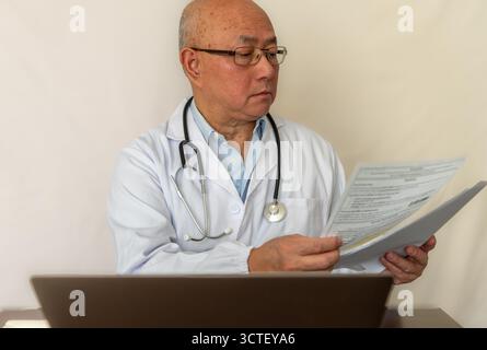 A doctor wearing a white coat reading medical documents and working on a laptop computer on a desk. Stock Photo