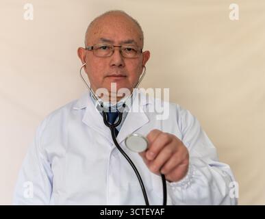 A doctor in a white coat using a stethoscope against a beige background. Stock Photo