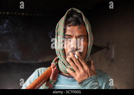 Dhaka, Bangladesh - 24 January 2021: View of a man with a scarf on his head, exhaling smoke from a cigarette, a wooden stick resting on his shoulder. Stock Photo