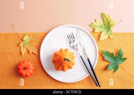 Thanksgiving arrangement with autumn table setting, yellow pumpkin, white plate and autumnal leaves on beige background. Top view, flat lay. Stock Photo