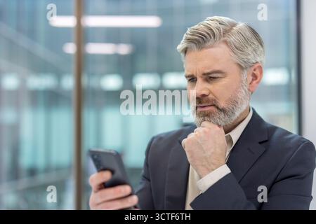 Mature corporate executive in a suit intently using a smartphone in a modern office, thinking and planning business strategy while checking messages and data for solutions Stock Photo