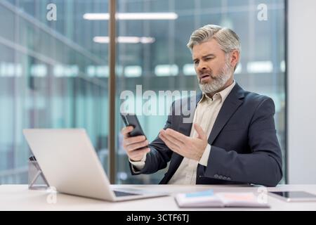 Senior businessman in suit at desk reacting with shock and frustration to bad news on his smartphone, gripping device while laptop sits open in modern office environment Stock Photo