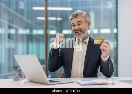 Energetic senior businessman sitting at his office desk, holding a credit card and raising his fist in celebration while actively engaging in online shopping using a laptop Stock Photo