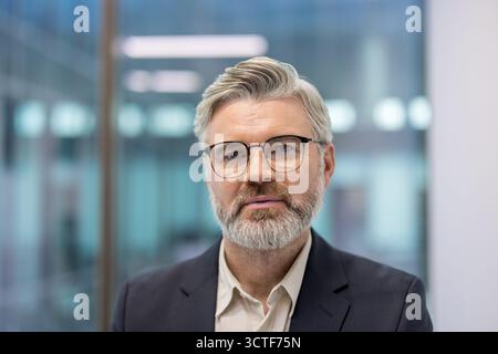 Senior businessman standing confidently in a modern office environment, wearing glasses and a suit, conveying professionalism, experience, and leadership in a corporate setting Stock Photo