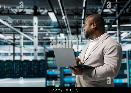 Programmer in data center using laptop, reading error logs, doing checkup to prolong hardware life span and avoid downtime. Server hub workplace employee monitoring infrastructure rigs using notebook Stock Photo