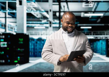 Happy data center engineer writing lines of code running on tablet, troubleshooting rackmounts. Cheerful IT professional using programming scripts on device in server room Stock Photo
