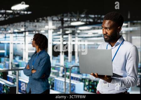Smiling black technician and colleague in data center reviewing large language model visualizations on PC. Professional IT engineering team analyzing neural network performance outputs. Stock Photo