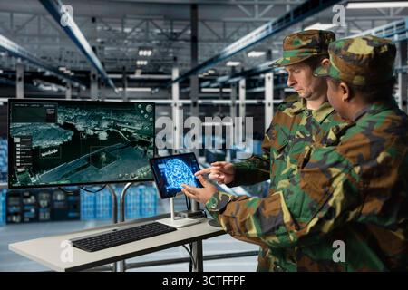 Military experts in data center reviewing drone surveillance feed using AI on tablet. Army units in high tech server room doing battlefield reconnaissance operations using artificial intelligence Stock Photo