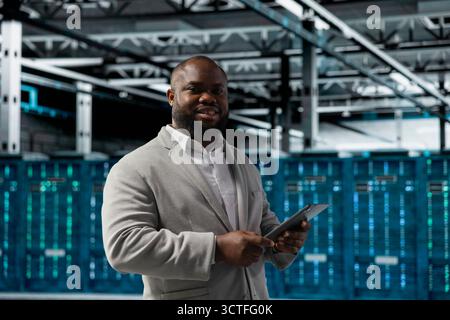 Portrait of cheerful technician in server hub checking recovery plan on tablet, monitoring energy consumption. Jolly data center staff member uses device, making sure sensors are functioning optimally Stock Photo