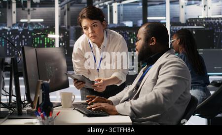Server room manager supervise technician examining deep learning algorithms. Data center team leader and African american employee review performance dashboards of AI equipment rigs, camera A Stock Photo