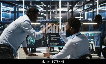 Server hub manager checking AI generated data analytics graphs, congratulating worker. Data center team leader doing fist bump with admin using artificial intelligence showing system metrics, camera A Stock Photo